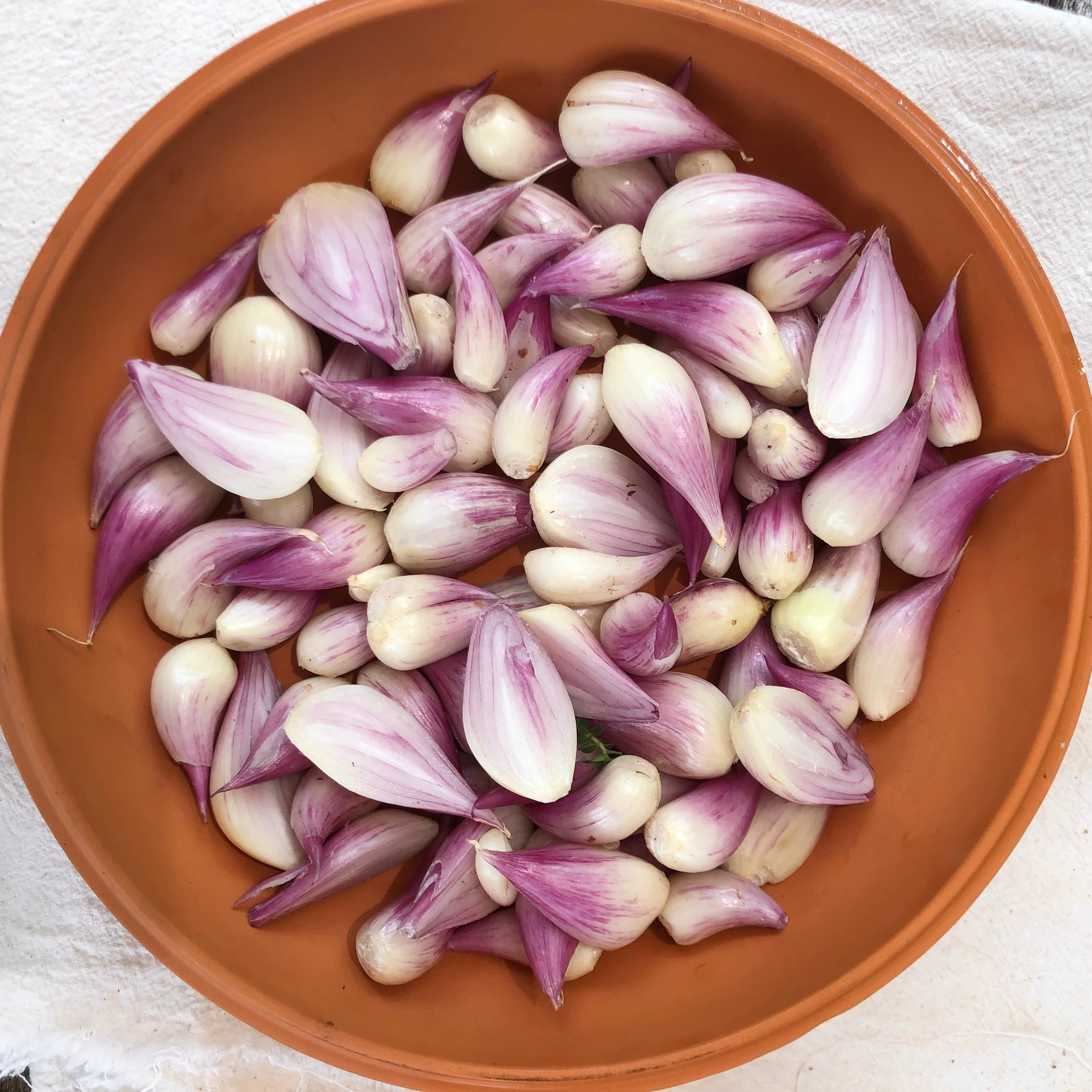 A top-down view of fresh shallot bulbs with purple and white skins inside a terracotta-colored bowl, set on a white textured fabric background.